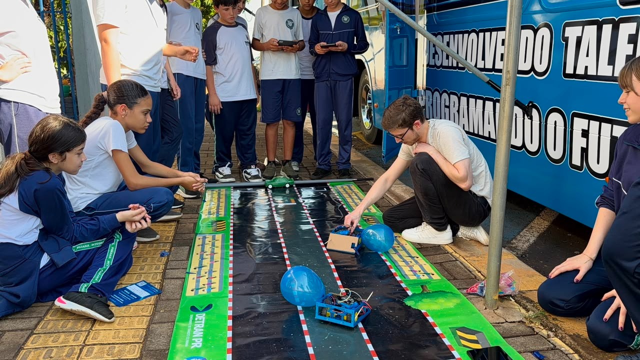 Alunos assistindo carrinhos-robô disputarem um desafio na pista montada ao lado do ônibus
